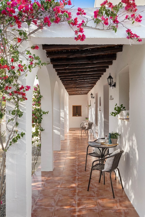 A covered outdoor area with a tiled floor, white walls, wooden beams, and potted plants.