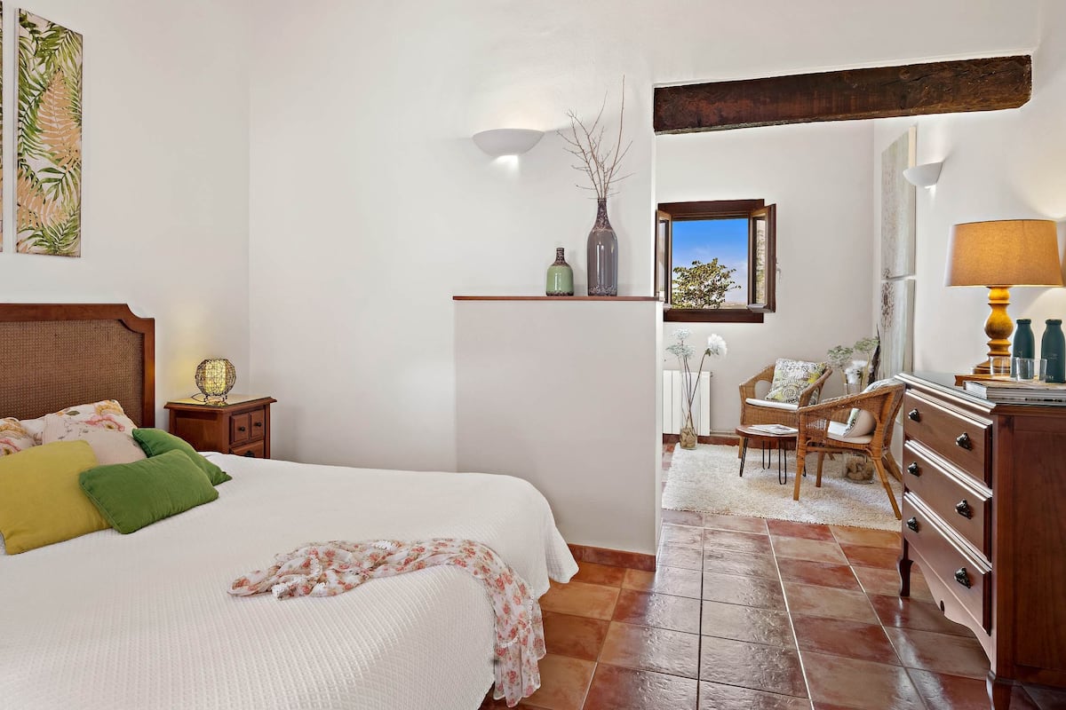 Um quarto com cama, mesa de cabeceira, cômoda, cadeira, janela e uma parede com obras de arte.