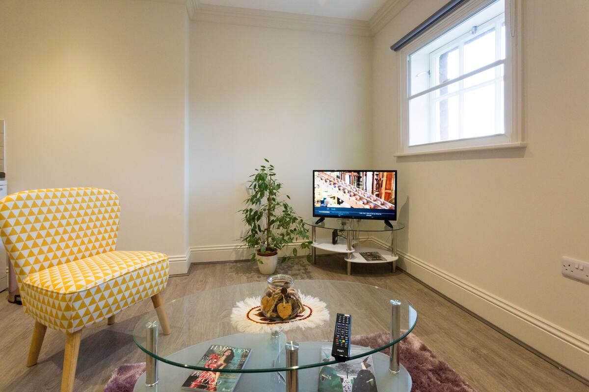 A living room with a yellow patterned armchair, a glass coffee table, a potted plant, and a television on a stand.