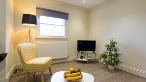 Un salón con un sillón estampado, una mesa redonda con fruta, un televisor y una planta en maceta.