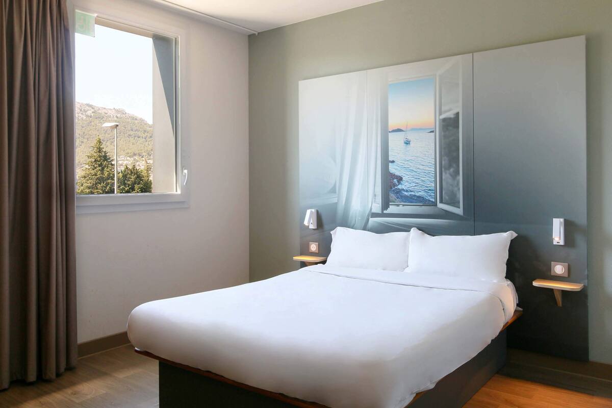 A neatly made bed in a hotel room with a large window, a painting of a coastal scene, and bedside tables with lamps.