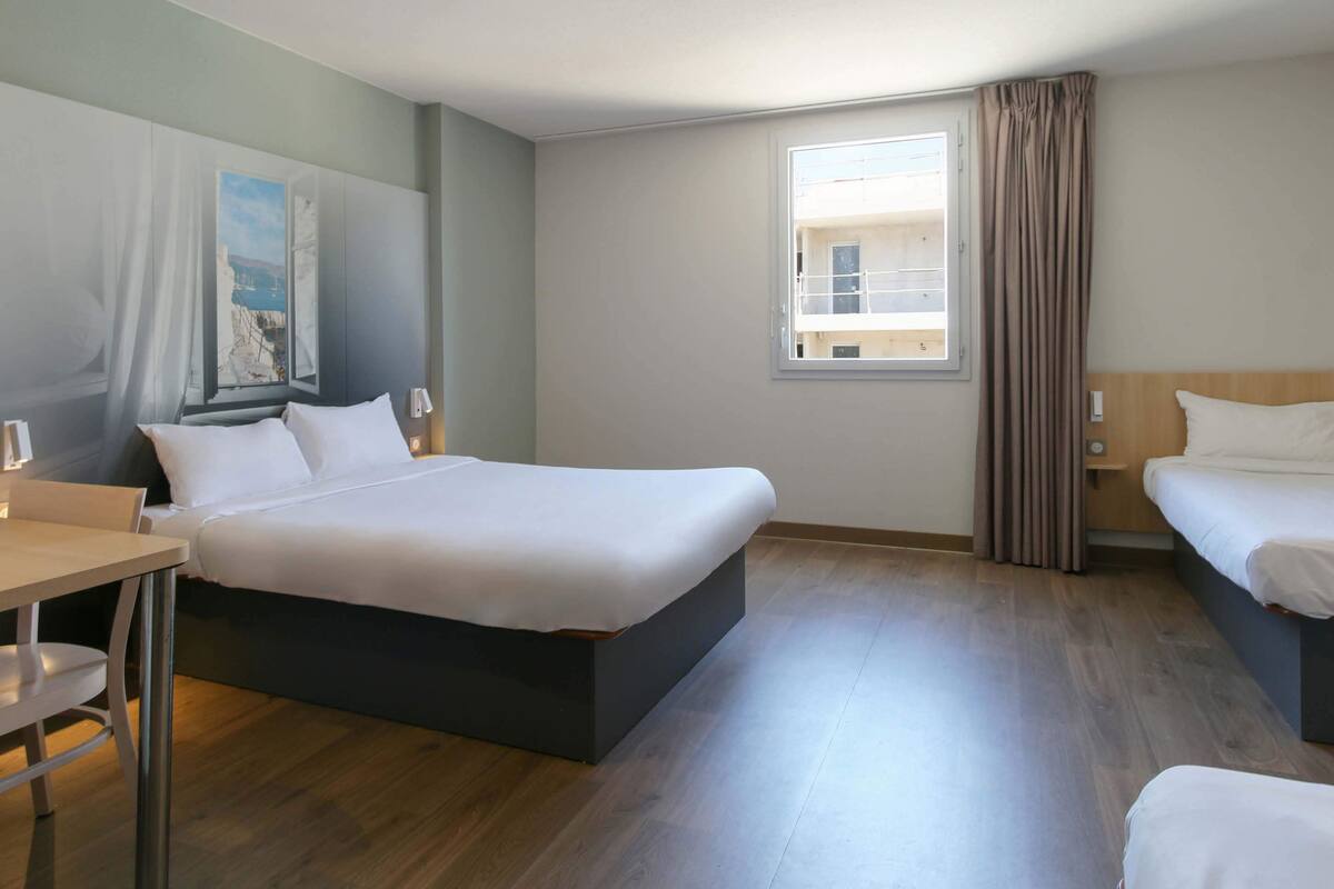 A hotel room with a large bed, a desk, a chair, a window with curtains, and a painting on the wall.