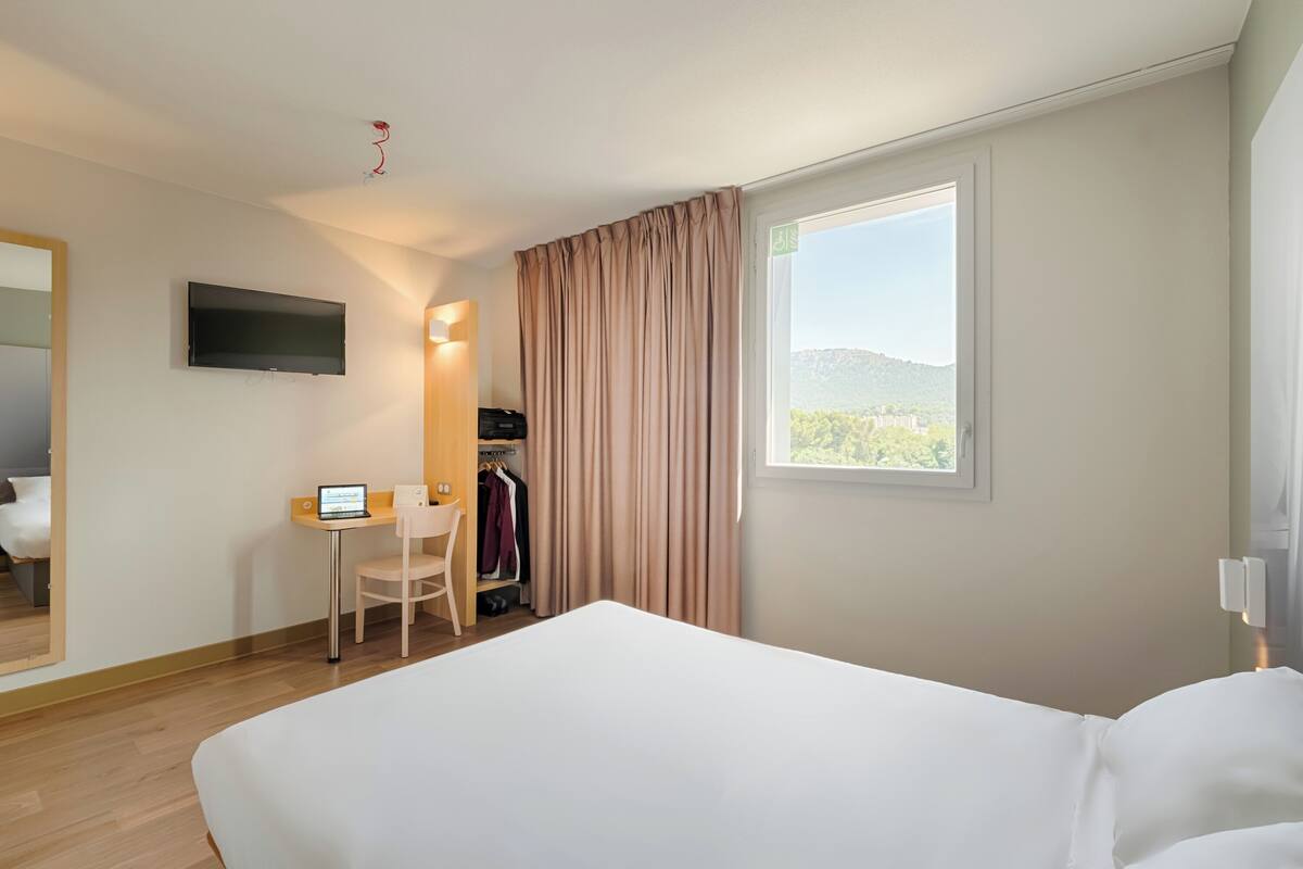 A hotel room with a bed, a desk with a laptop, a TV, and a window with curtains.