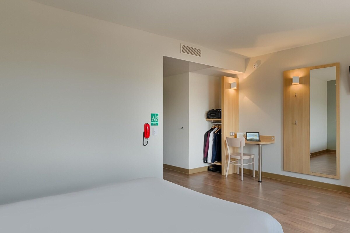 Quarto de hotel com cama, escrivaninha com laptop, cabideiro e espelho.
