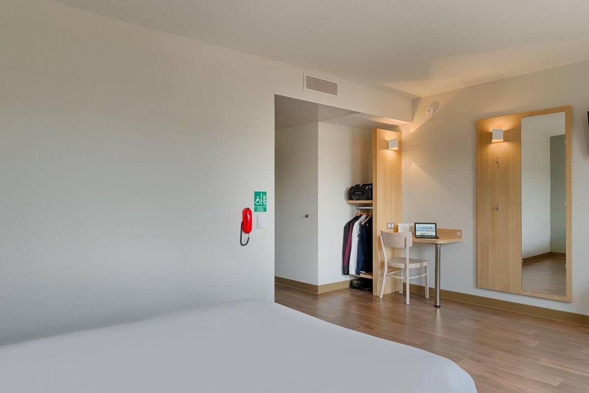 A hotel room with a bed, a desk with a laptop, a coat rack, and a mirror.