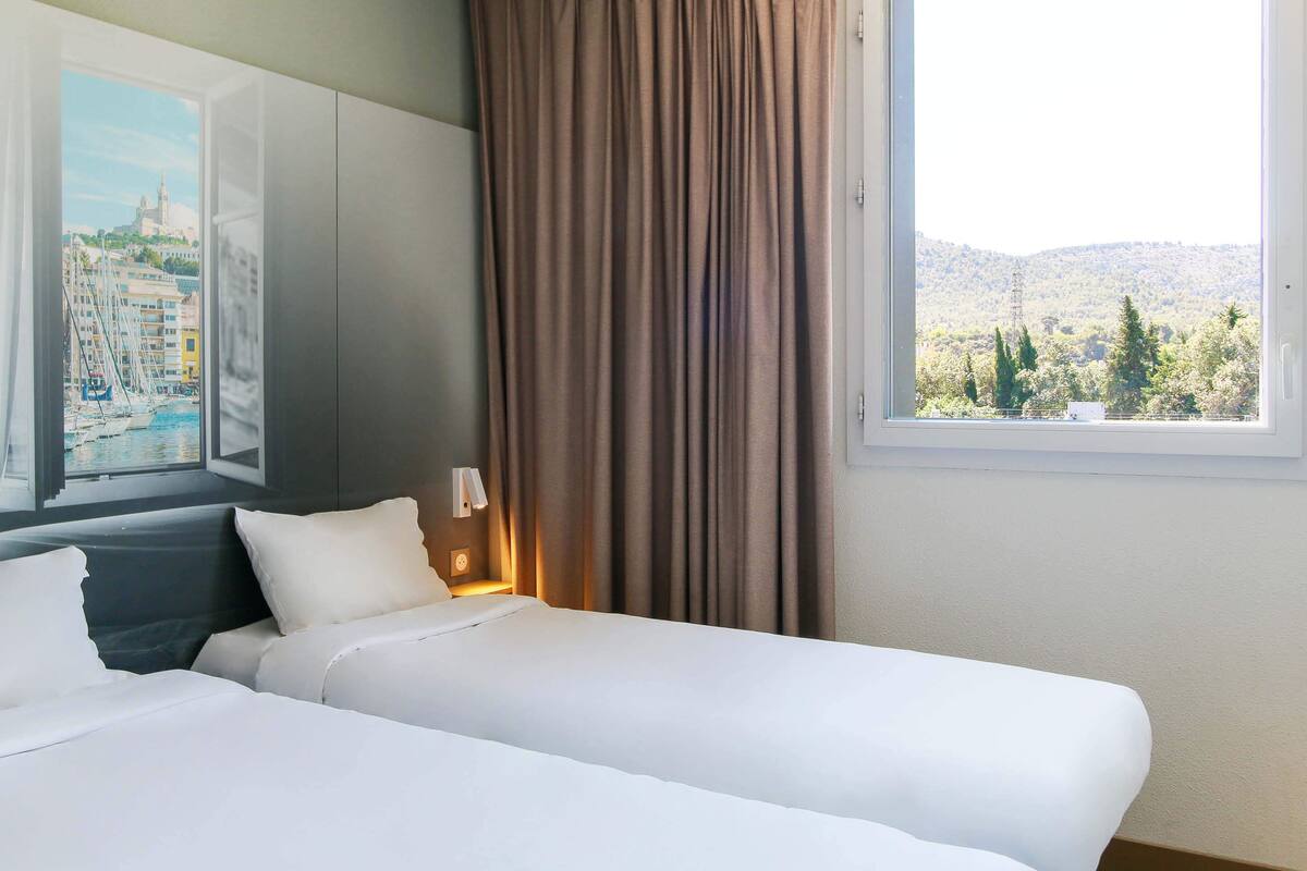 A hotel room with two beds, a nightstand with a lamp, a window with a view of a cityscape and hills, and a painting on the wall.