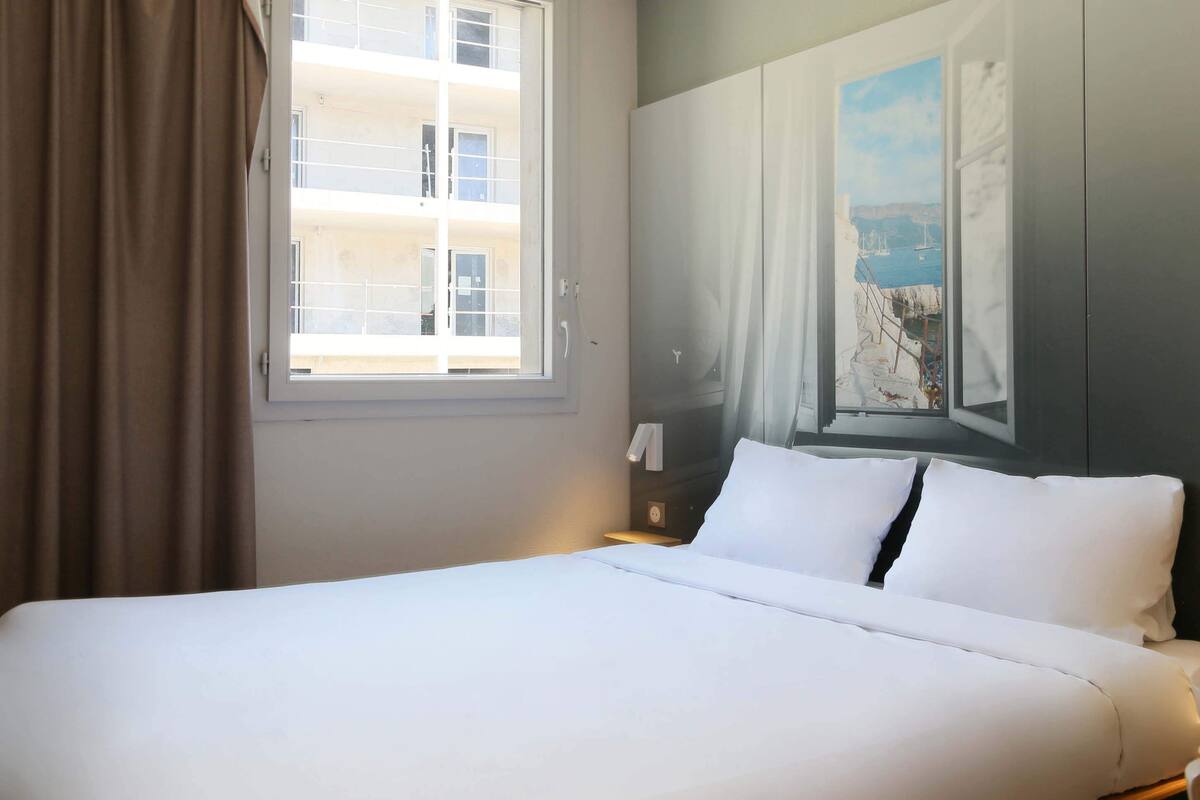A hotel room with a bed, a window with curtains, a nightstand, and a painting on the wall.