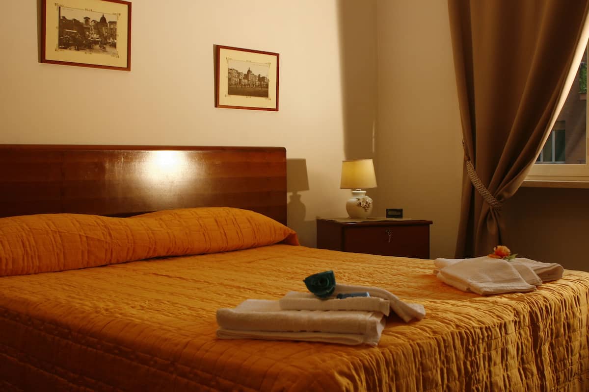 Una cama bien tendida con cabecera de madera, dos cuadros enmarcados en la pared, una lámpara de noche y una ventana con cortinas.