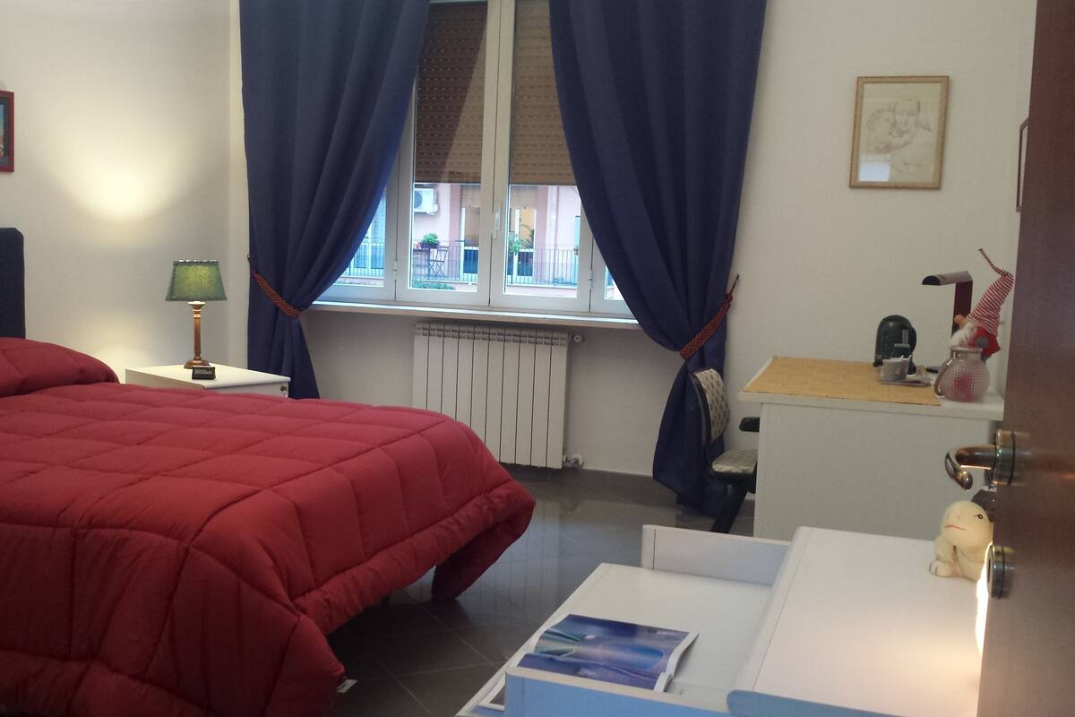 Un dormitorio con cama, mesita de noche, lámpara, ventana con cortinas, radiador y escritorio con silla.