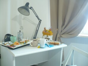 Un escritorio con una lámpara, una cafetera, una taza, un plato con comida y una cesta de pan.
