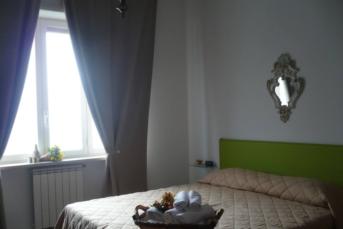 Una cama bien hecha con una colcha acolchada, una mesita de noche con una taza y una planta pequeña, y una ventana con cortinas.