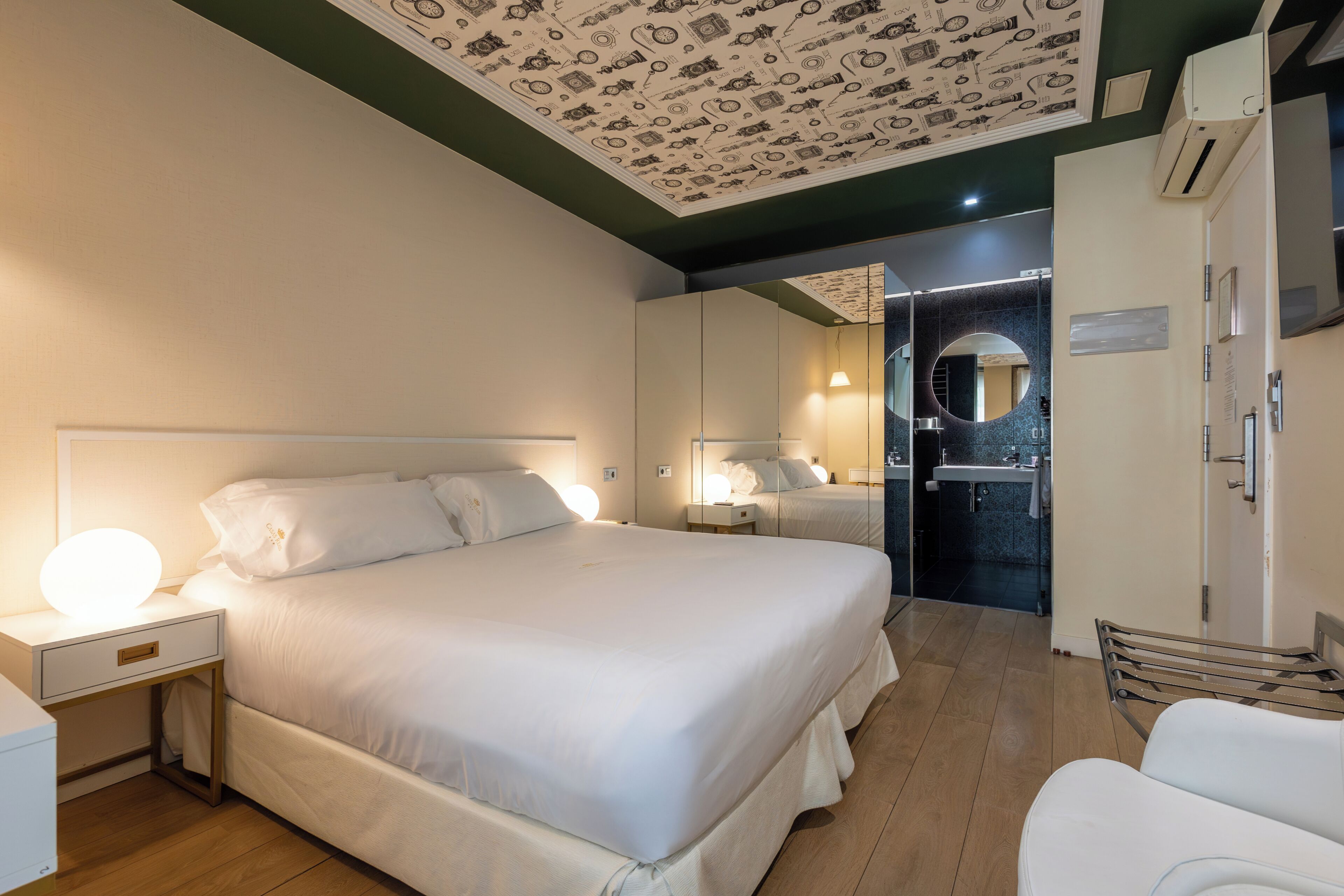 A hotel room with a large bed, bedside table with a lamp, a mirror, a wardrobe, and a bathroom visible through an open door.