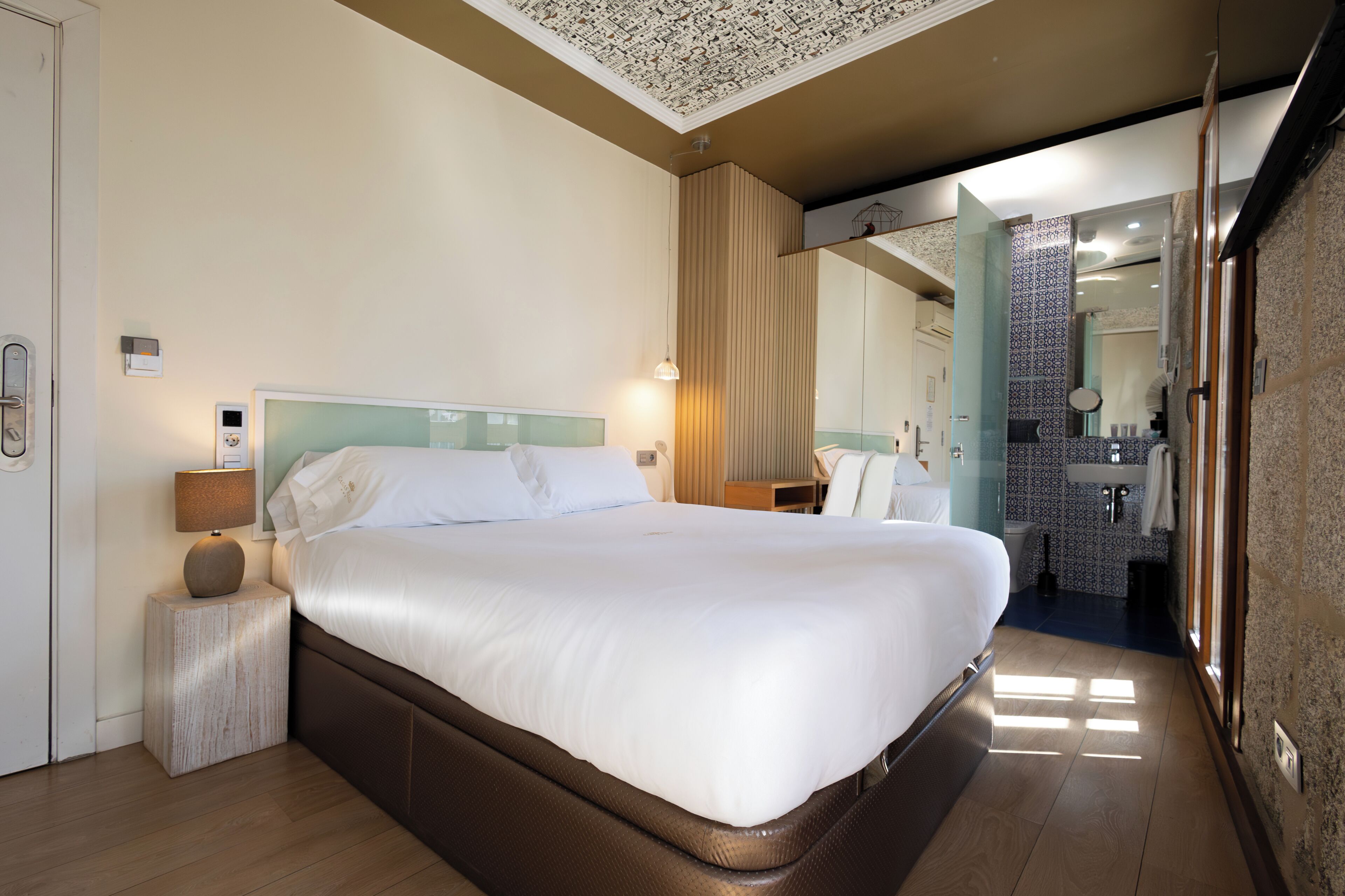 A hotel room with a large bed, bedside table, lamp, and a bathroom visible through an open door.