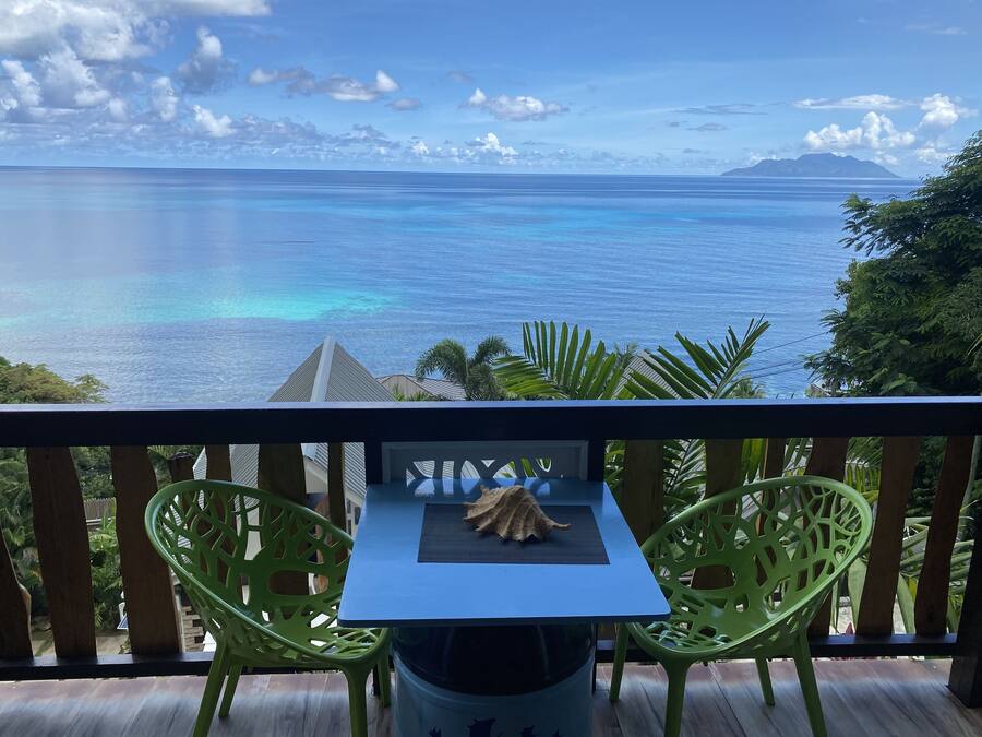 Un balcón con mesa y sillas frente a un mar azul y una cadena montañosa distante.