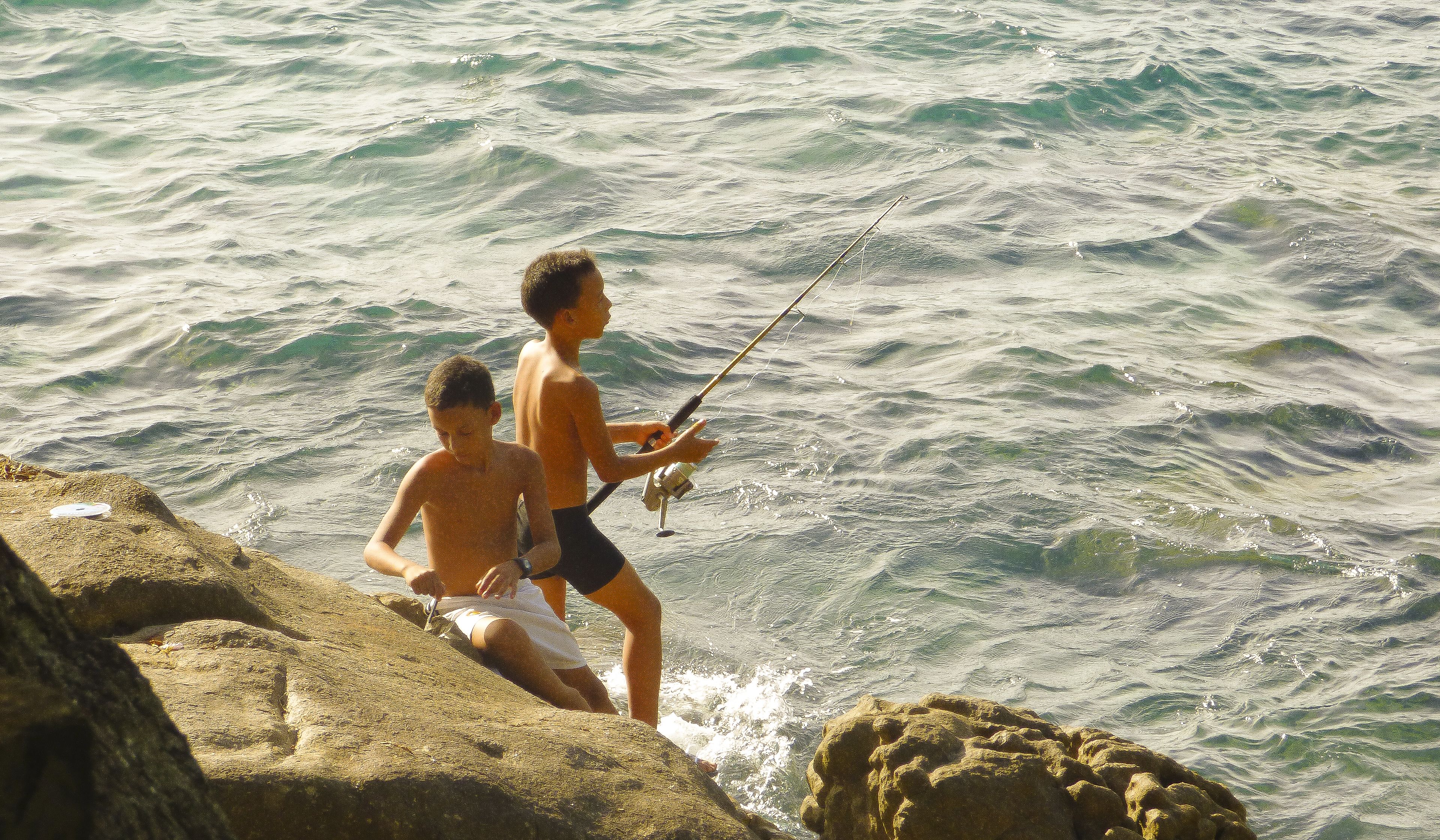 Due persone che pescano lungo una costa rocciosa, con le onde che si infrangono sullo sfondo.