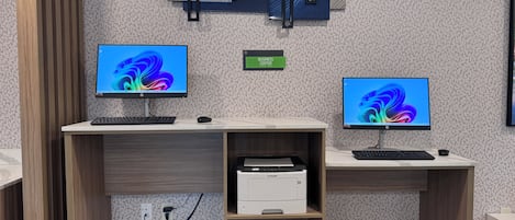 Ein moderner Arbeitsbereich mit zwei Schreibtischen, jeweils ausgestattet mit Computerbildschirm, Tastatur und Maus. Ein Schild kennzeichnet den Businessbereich, und ein Drucker ist auf einem der Schreibtische zu sehen.