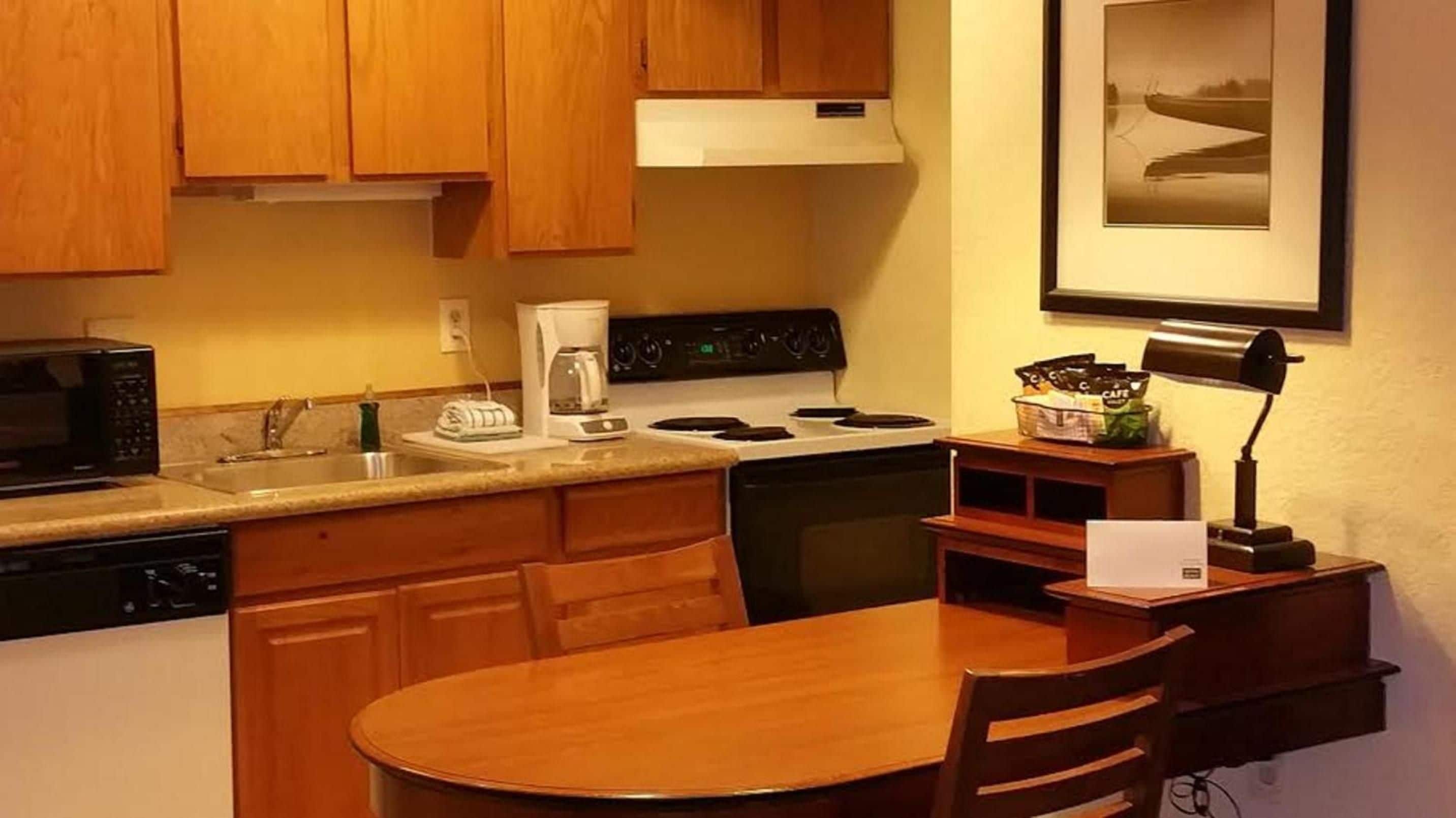 A compact kitchen with wooden cabinets, a microwave, a coffee maker, and a stove. There is a dining table with chairs and a framed picture on the wall.