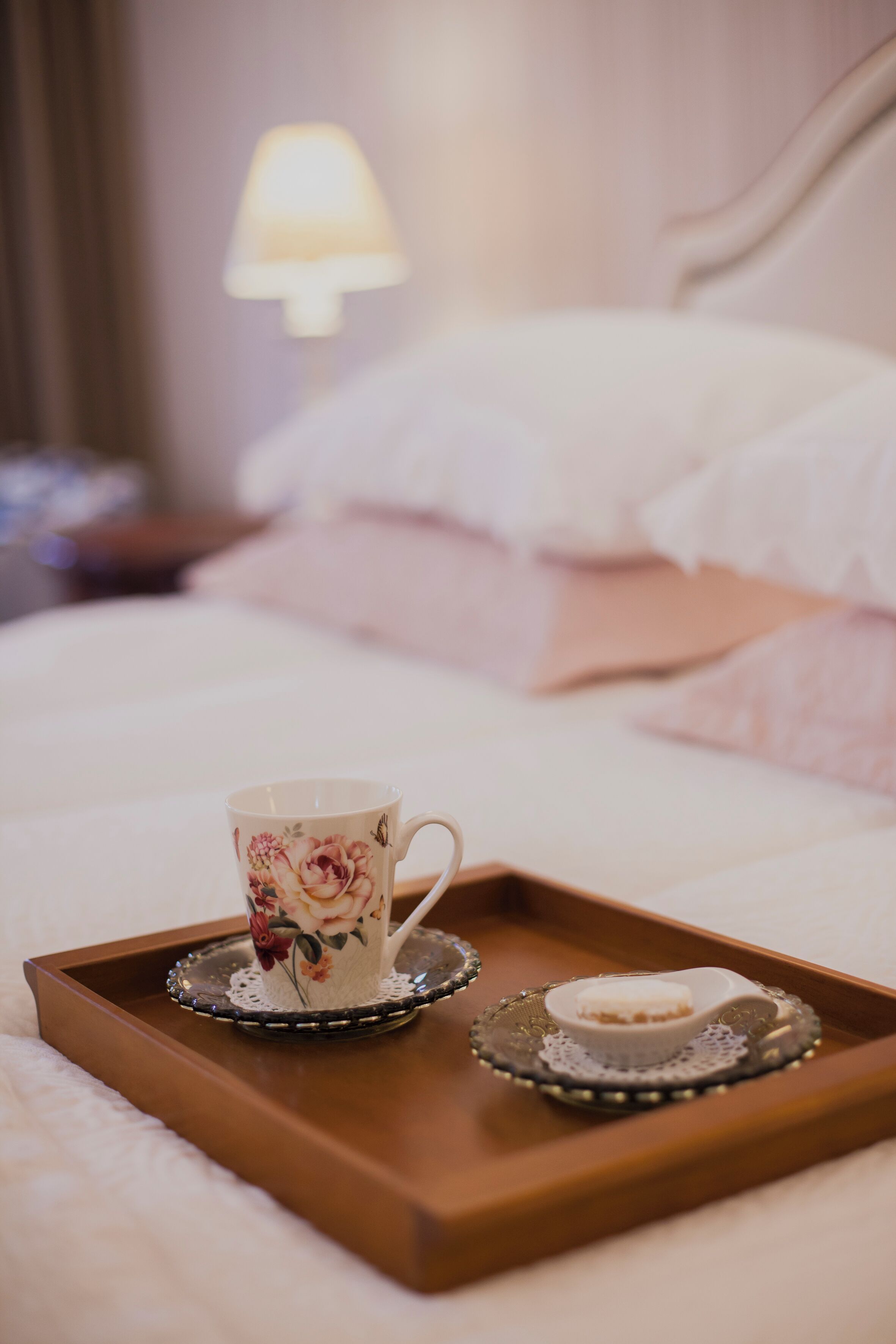 Ein Tablett mit einer Blumen-Mug und einer kleinen Schale auf einem Bett mit weißer Bettwäsche, im Hintergrund eine Lampe.
