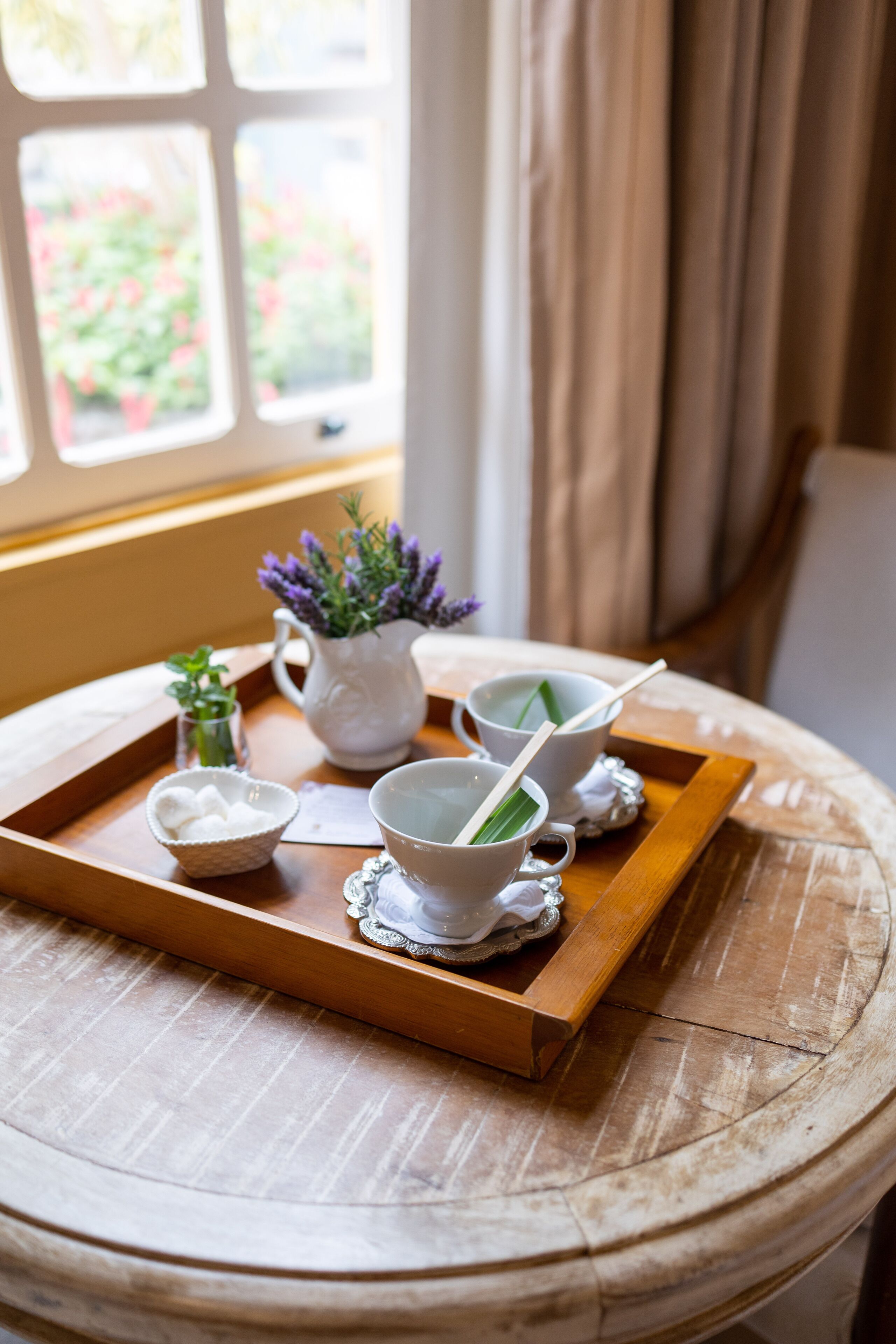 A wooden tray with teacups, a pitcher, and a small bowl on a round table near a window with curtains.