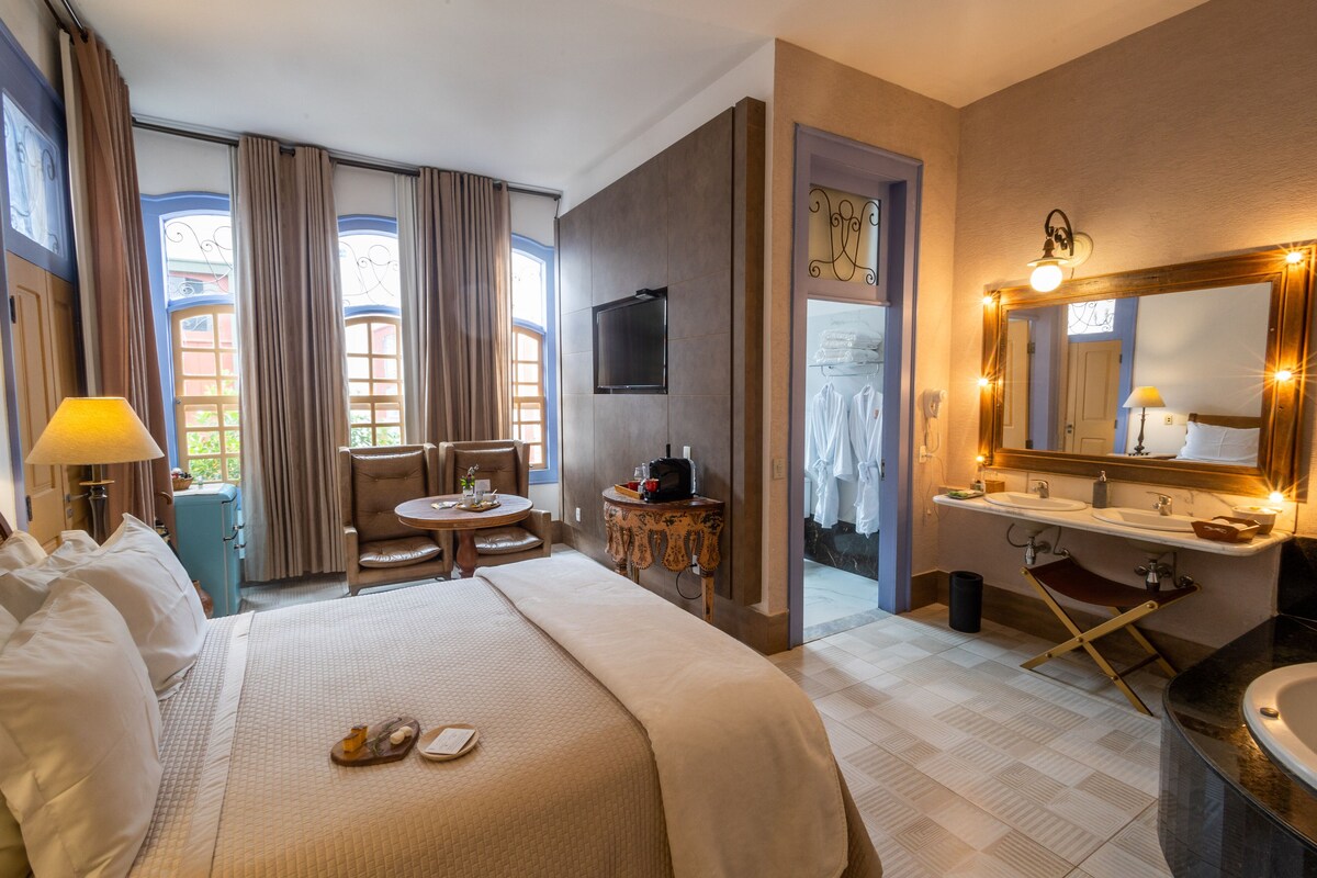 A hotel room with a bed, a desk, a chair, a TV, a bathroom with a sink and mirror, and a window with curtains.
