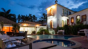 A pool area with lounge chairs and towels, a well-lit building with a balcony, and a clear sky. - Pousada Pequena Tiradentes (Tiradentes)
