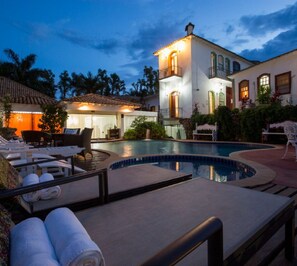 A pool area with lounge chairs and towels, a well-lit building with a balcony, and a clear sky. - Pousada Pequena Tiradentes (Tiradentes)