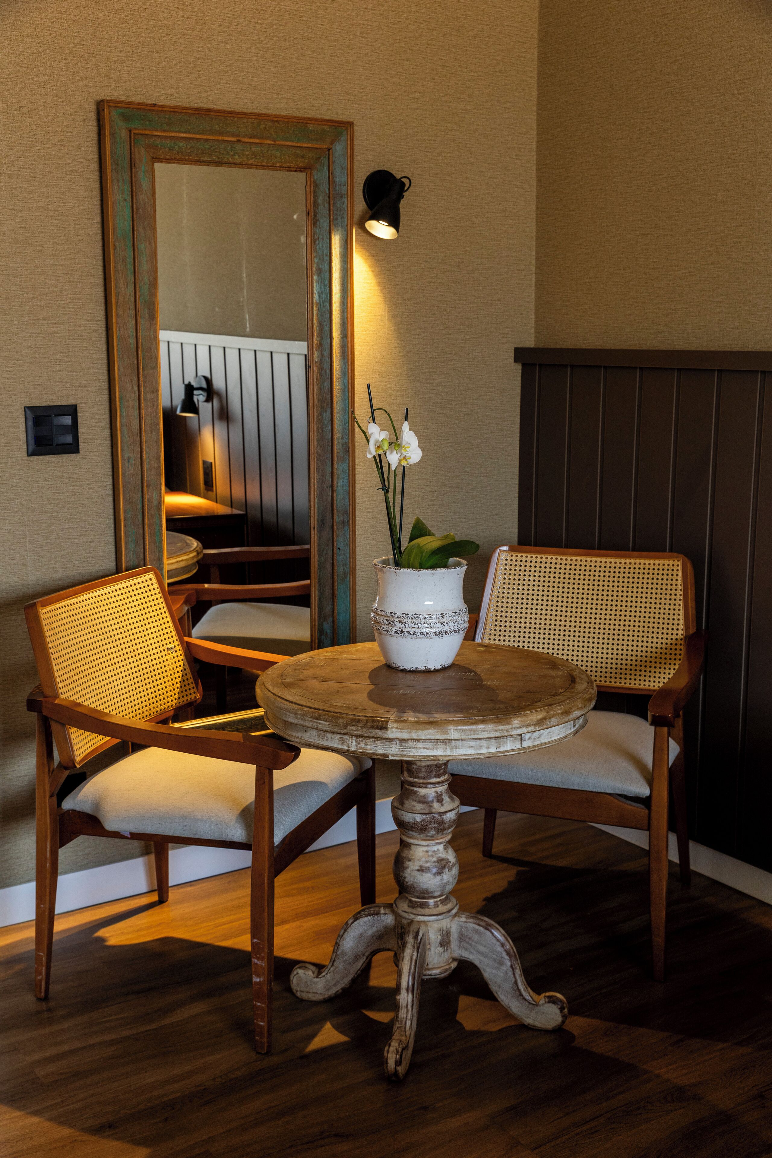 A dining area with a round wooden table, two wicker chairs, a white vase with flowers, and a mirror on the wall.