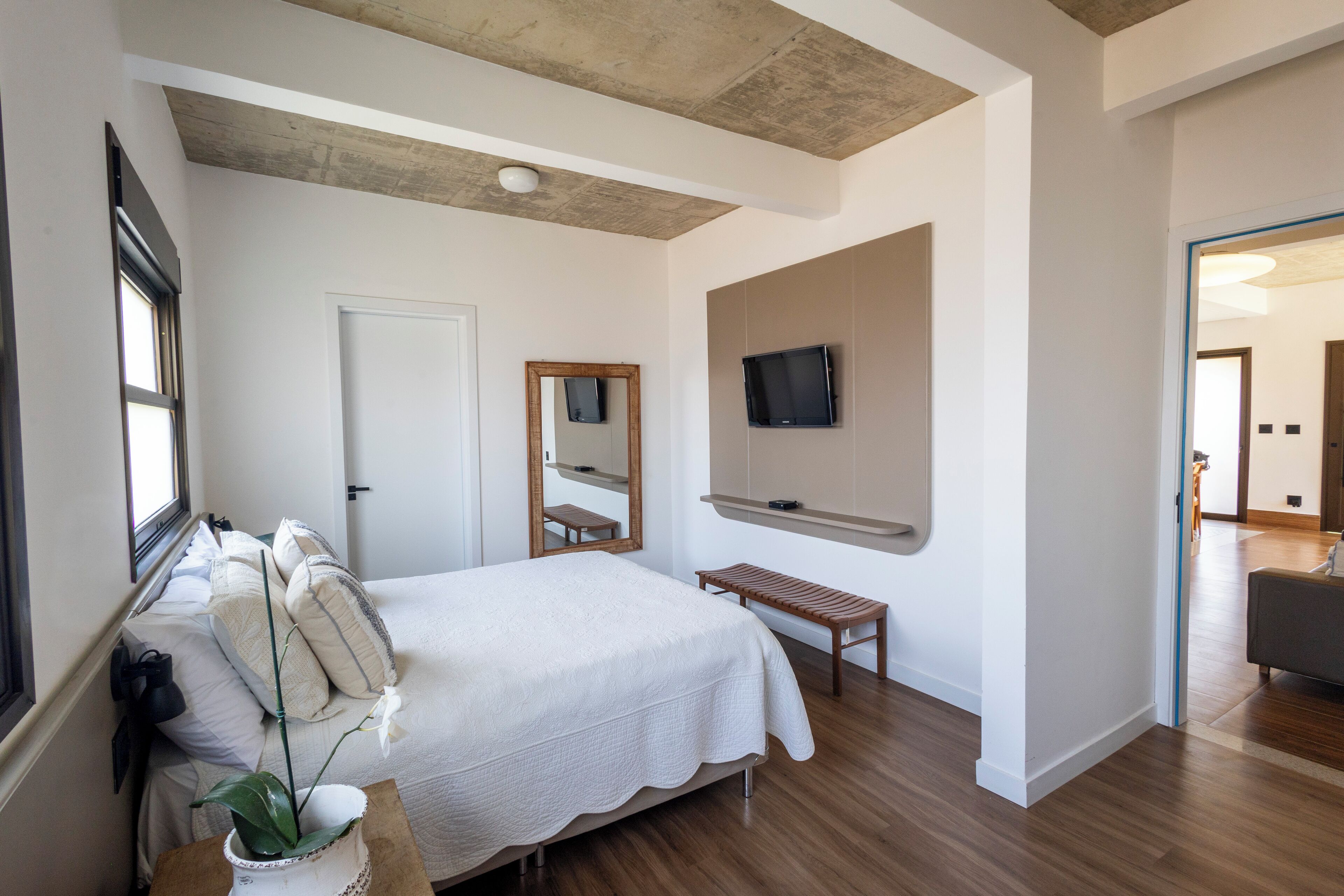 A bedroom with a bed, a wooden bench, a mirror, and a television on the wall.
