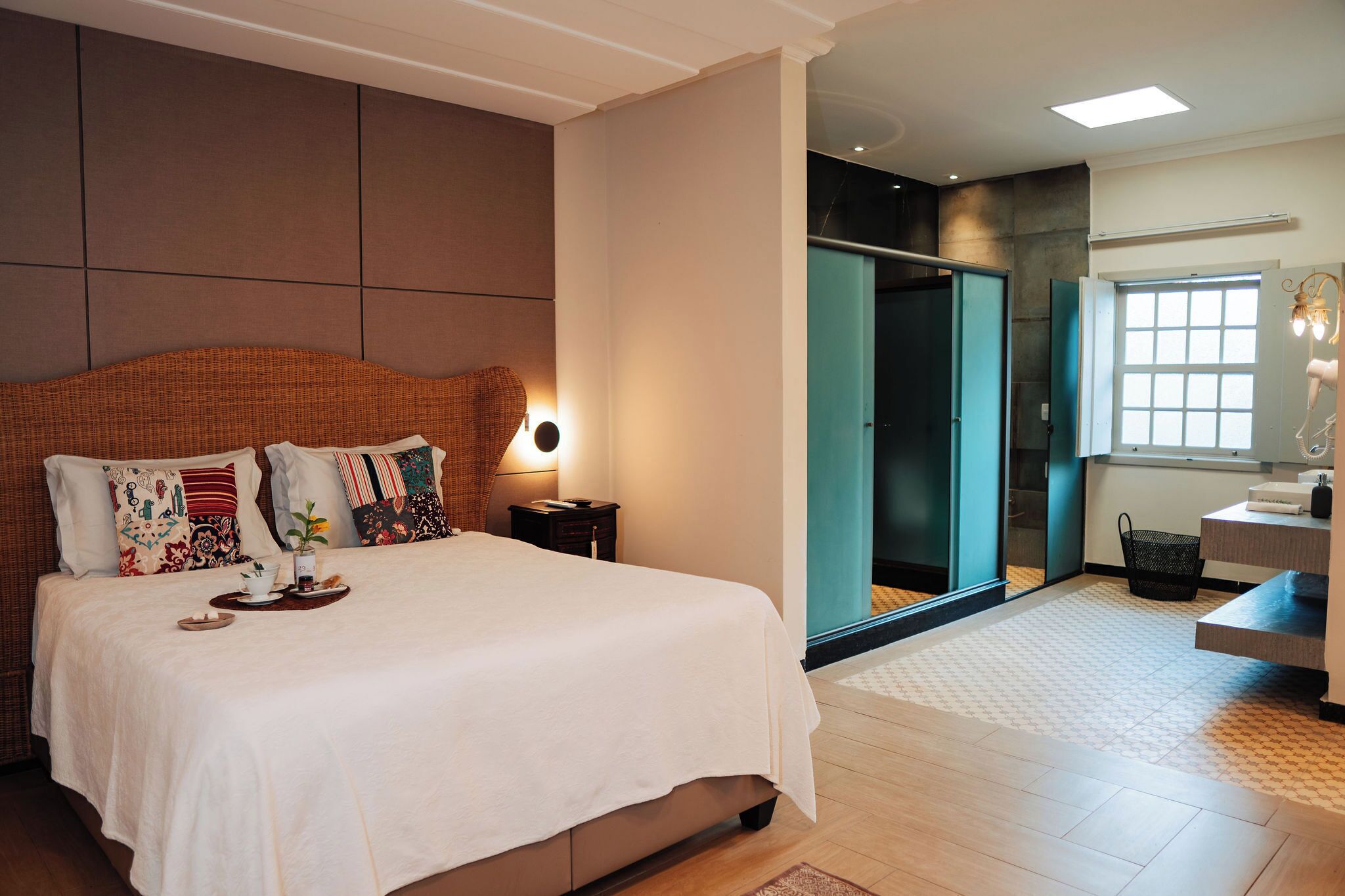 Quarto de hotel moderno com uma cama grande, uma mesa de cabeceira, um espelho, uma janela e uma porta para um banheiro.