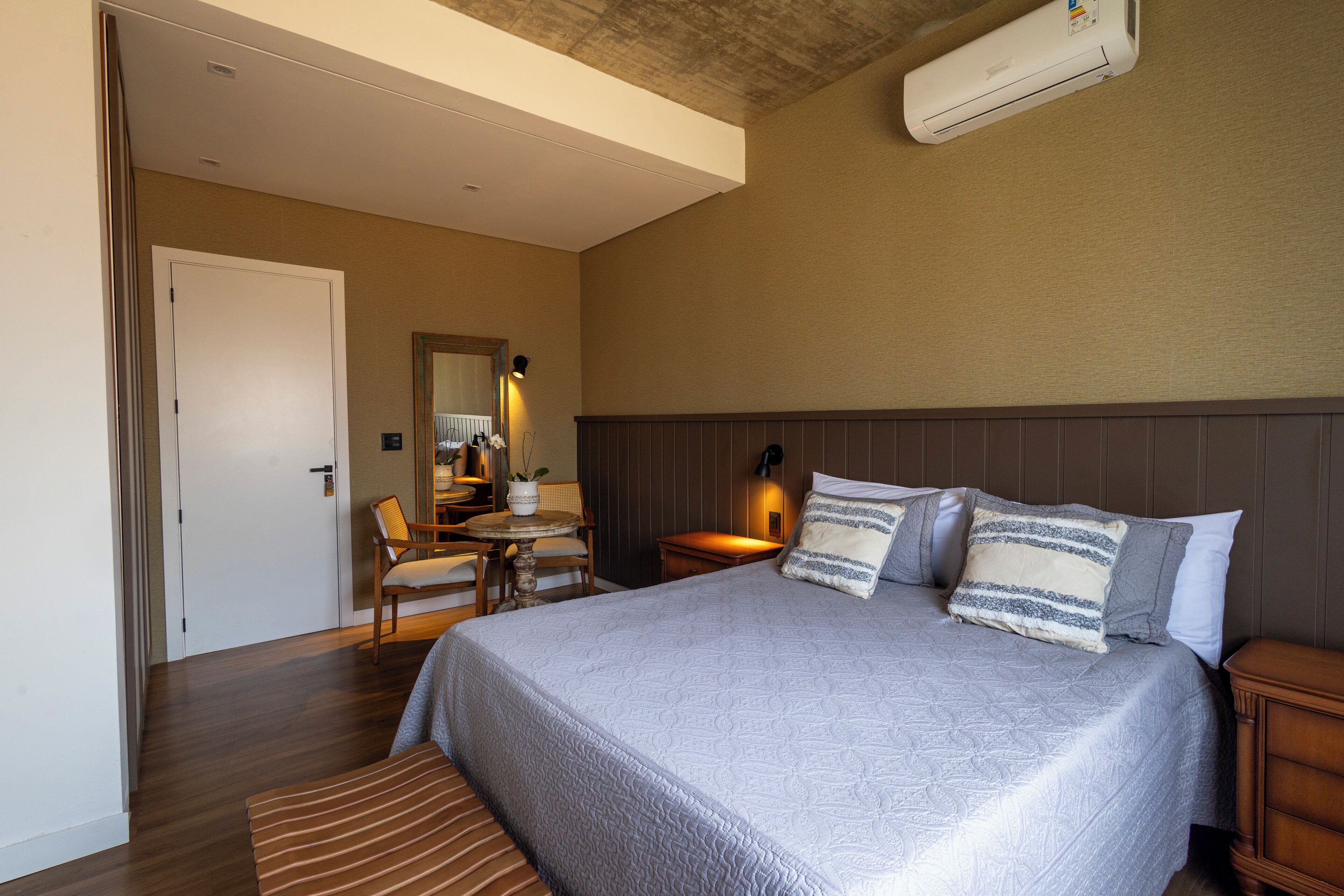 Habitación de hotel con una cama grande, una mesita de noche de madera, una pequeña mesa con sillas y un aire acondicionado instalado en el techo.