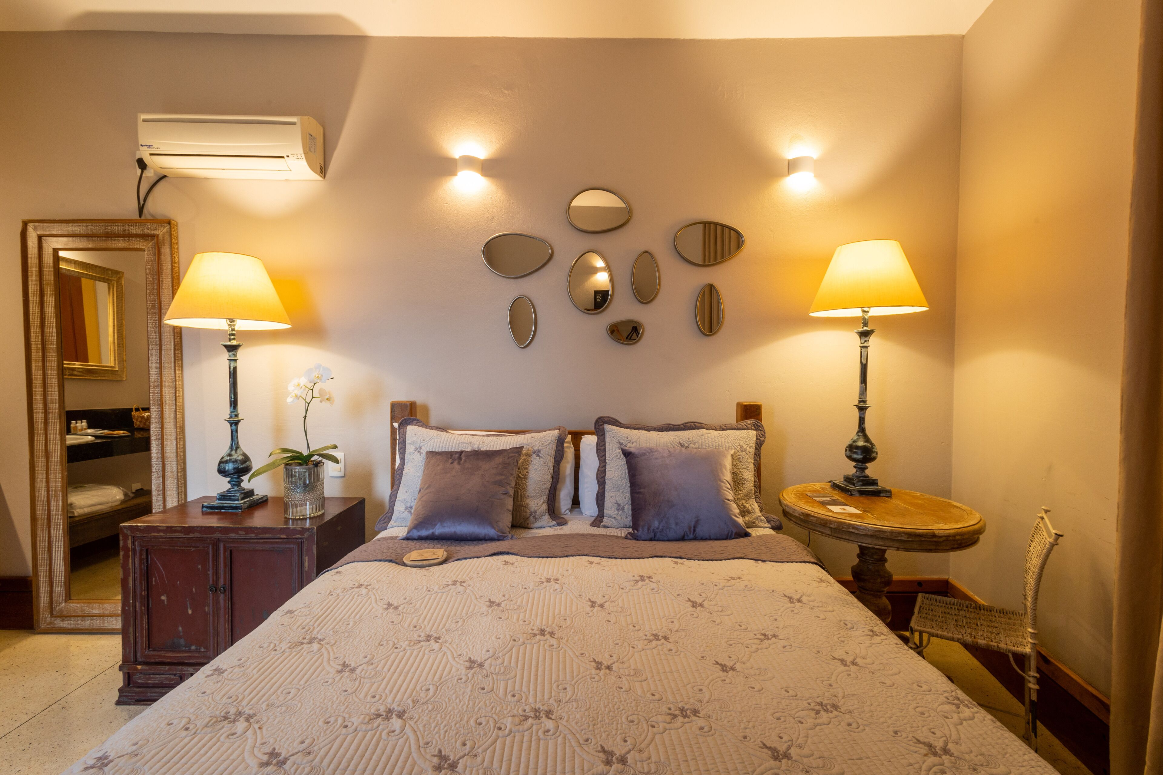 Un dormitorio ordenado con cama, mesitas de noche, lámparas, un espejo e iluminación en la pared.