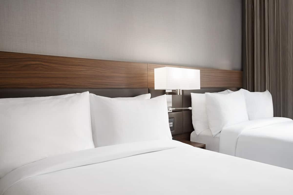 Una habitación de hotel con cabecera de madera, dos camas con sábanas blancas, una mesita de noche con una lámpara y vistas a una cortina.