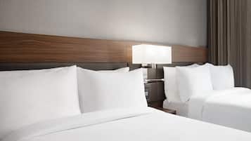 A hotel room with a wooden headboard, two beds with white linens, a bedside table with a lamp, and a view of a curtain.
