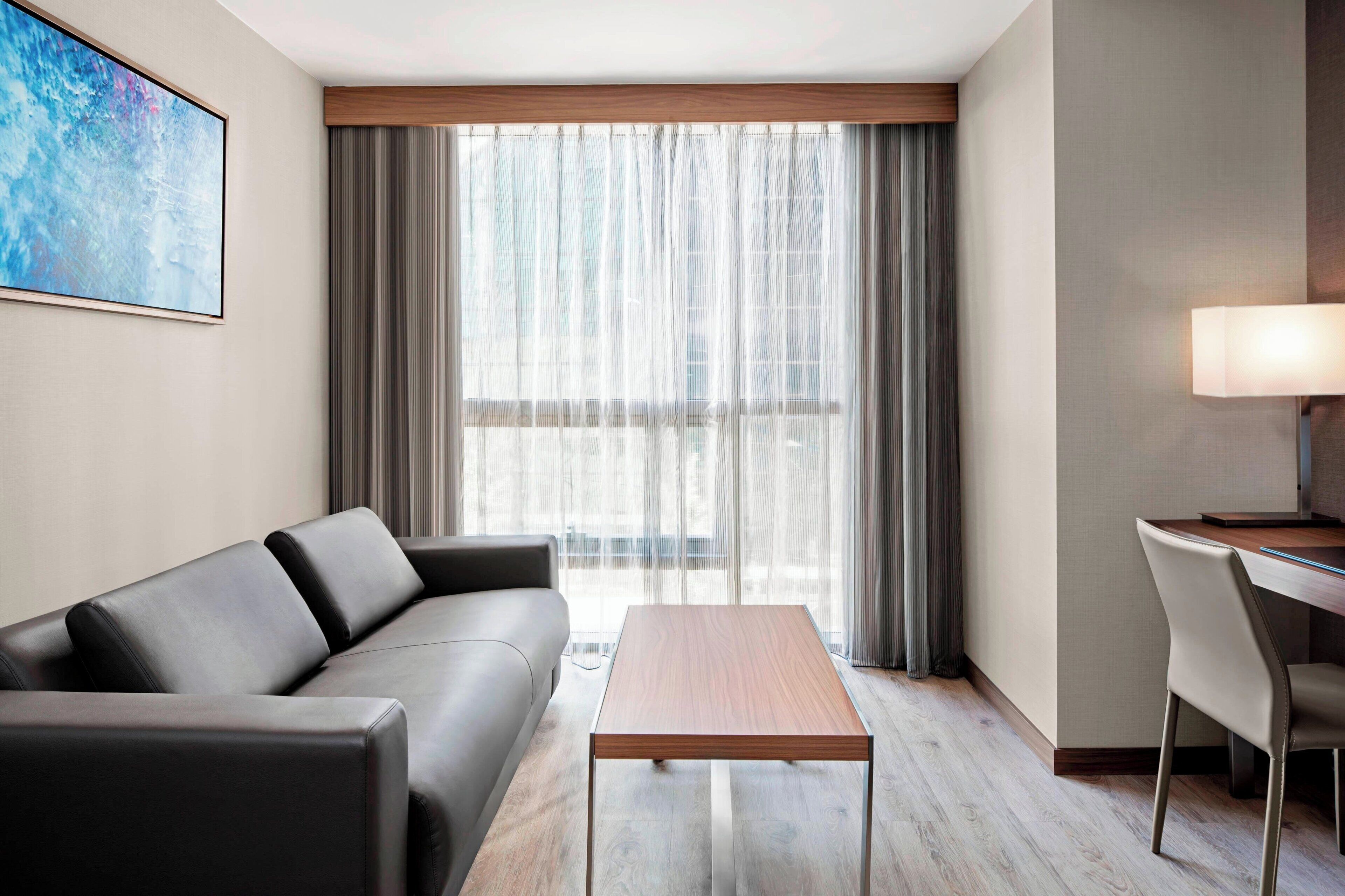 A modern hotel room with a leather sofa, a wooden coffee table, a desk with a lamp, and a large window with sheer curtains.
