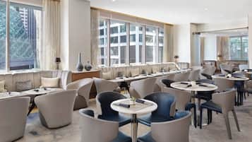 A modern hotel lobby with a long reception desk, multiple seating areas with round tables and chairs, large windows with curtains, and a view of the cityscape.