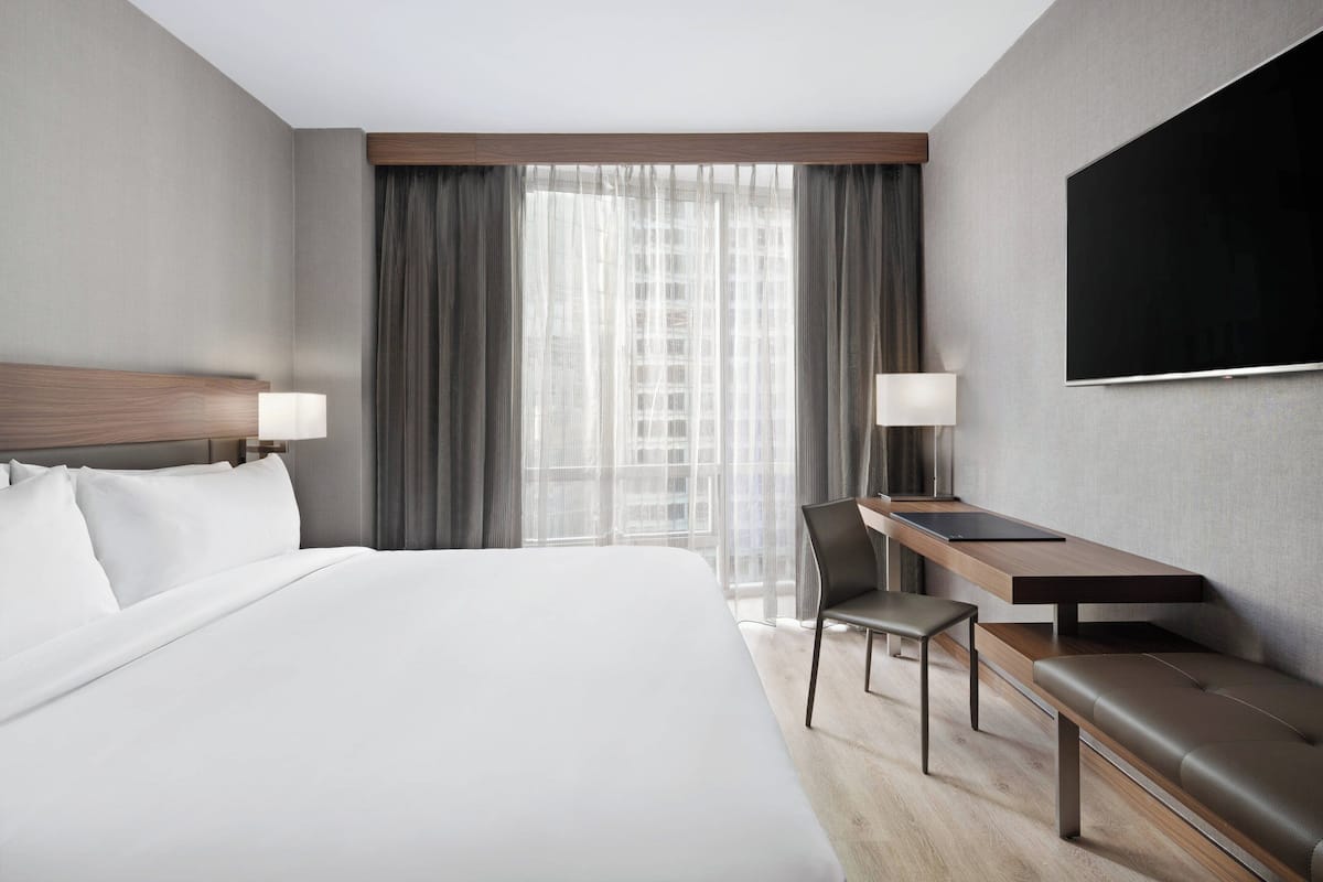 Habitación de hotel con una cama grande, un escritorio con silla, un banco, un televisor y una ventana con cortinas.