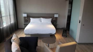 A modern hotel room with a large bed, bedside tables, a small round table, a sofa, and a view of the outside through a window.