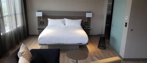 A modern hotel room with a large bed, bedside tables, a small round table, a sofa, and a view of the outside through a window.