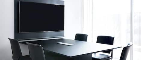 A conference room with a large flat-screen TV mounted on the wall, a dark-colored table, and four black chairs with mesh backrests.