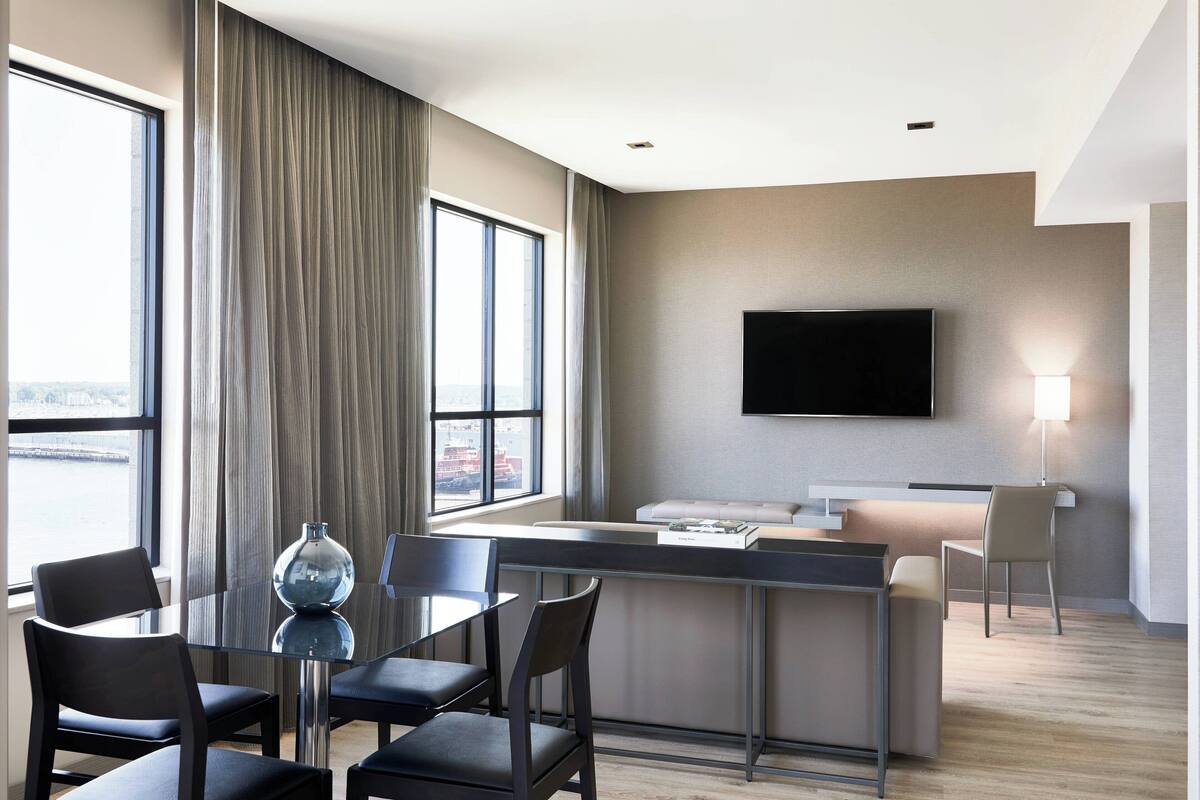 A modern hotel room with a glass dining table, black chairs, a sofa, a TV mounted on the wall, and a large window with curtains.