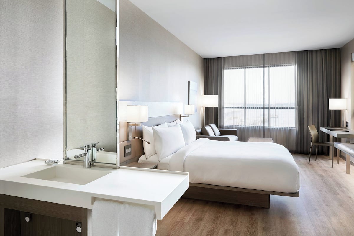 A modern hotel room with a large bed, a separate desk, a bathroom with a sink and mirror, and a large window with curtains.