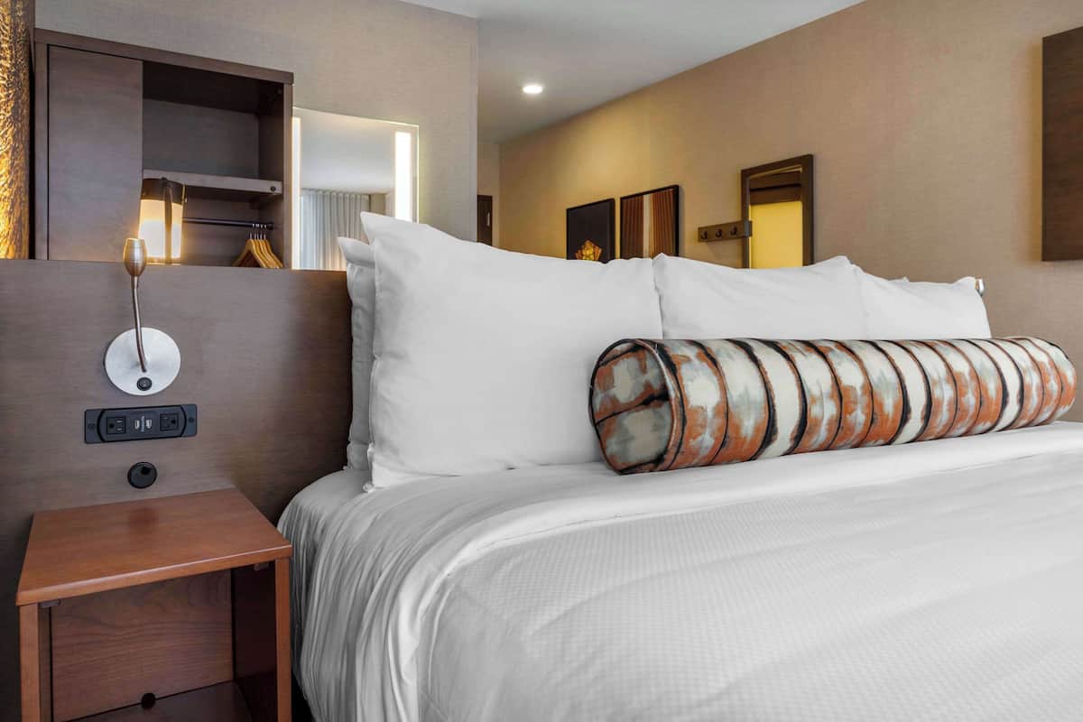 Ein Hotelzimmer mit einem ordentlich bezogenen Bett, einem Nachttisch mit Lampe, einer Steckdose und einem an der Wand befestigten Regal.