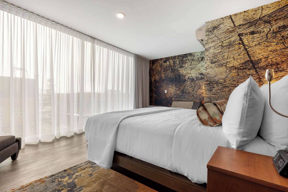 Ein modernes Hotelzimmer mit einem großen Bett, einem Nachttisch aus Holz und einem großen Fenster mit durchscheinenden Vorhängen.