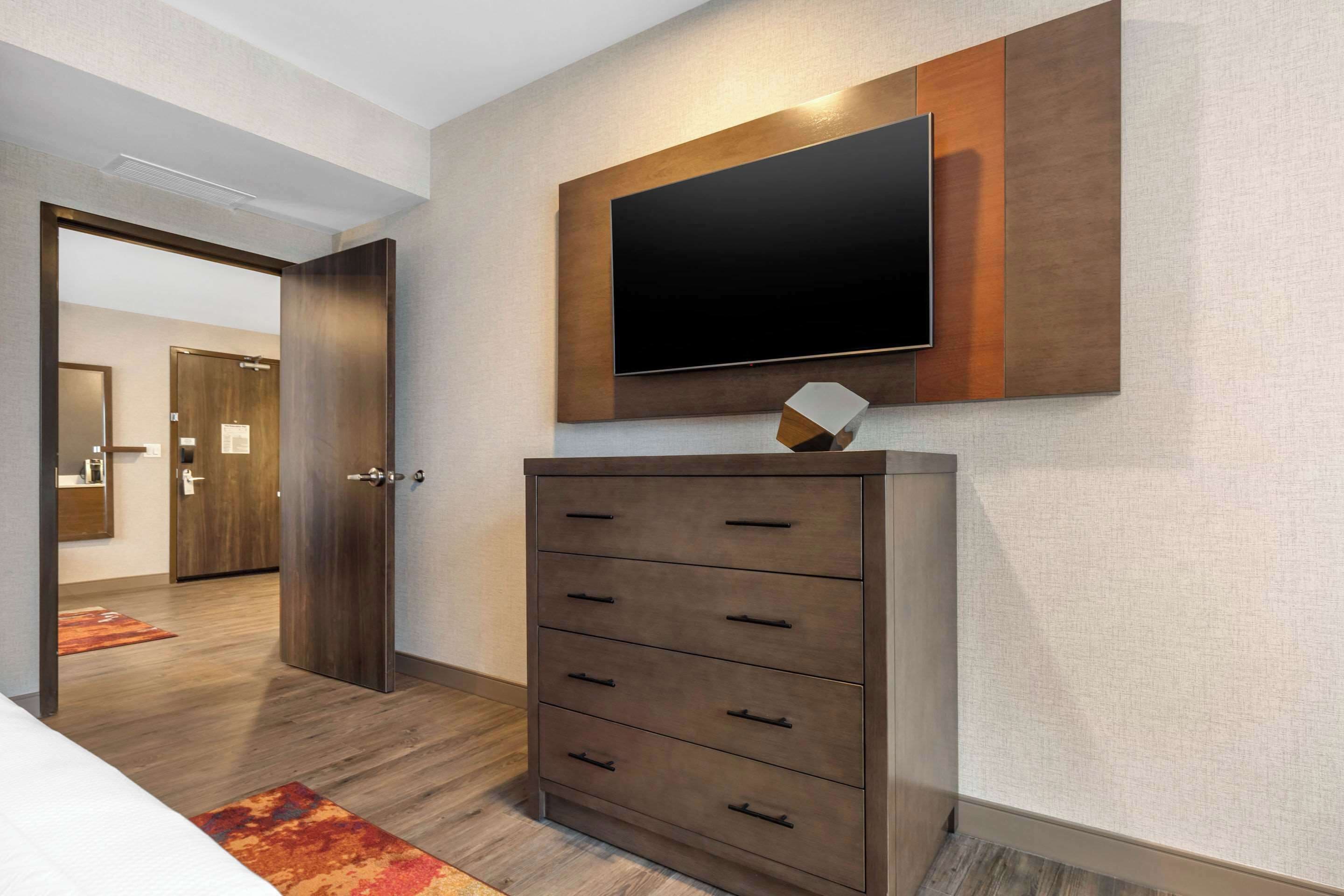 A hotel room with a large flat-screen TV mounted on the wall, a wooden dresser with drawers, and a carpeted floor.