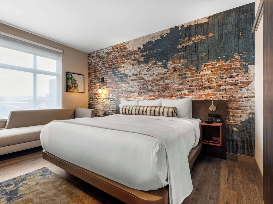 A modern bedroom with a large bed, a sofa, a brick wall, and a city view.
