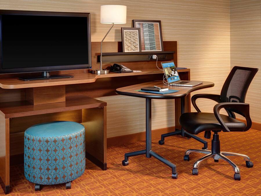 A hotel room with a wooden desk, a television, a lamp, a laptop, a chair, and a patterned ottoman.