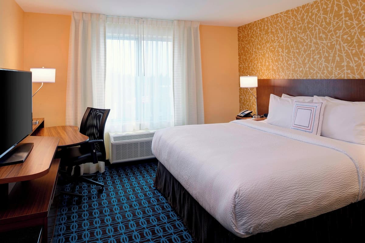 A hotel room with a bed, a desk with a TV, a chair, a window with curtains, and a patterned carpet.
