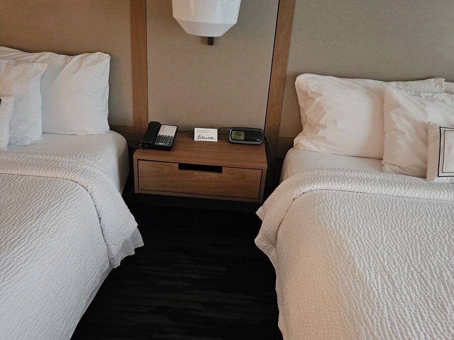 A hotel room with two beds, a bedside table with a lamp, a phone, and a small card.