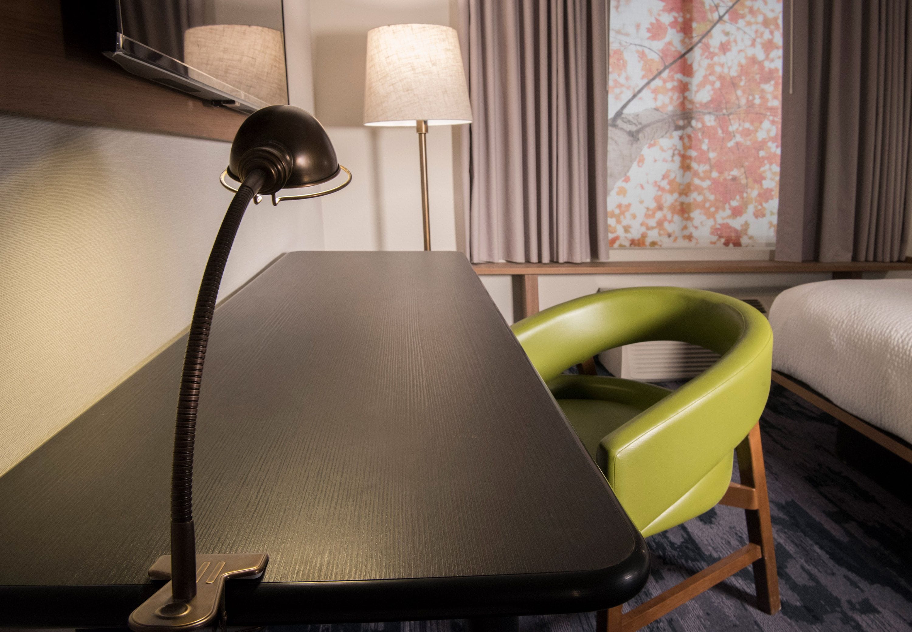 A hotel room with a desk, a green chair, a lamp, a bed, and a window with curtains.