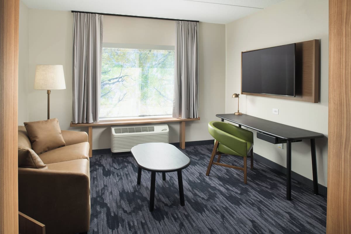 A hotel room with a sofa, a small table, a TV mounted on the wall, and a window with curtains.